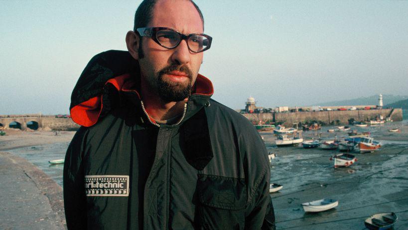 A younger Blue Juice director Carl Prechezer, who is in a green waterproof jacket with glasses on. Behind him is harbour. The boats are on the sand as the tide is out.