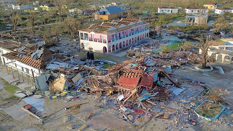 An aerial view of hurricane destruction shows collapsed buildings with roofs torn off and debris scattered across the ground. In the centre is a partially standing two-storey pink and white building. Broken wooden beams, metal sheets and concrete rubble cover the surrounding area as well as bare trees.