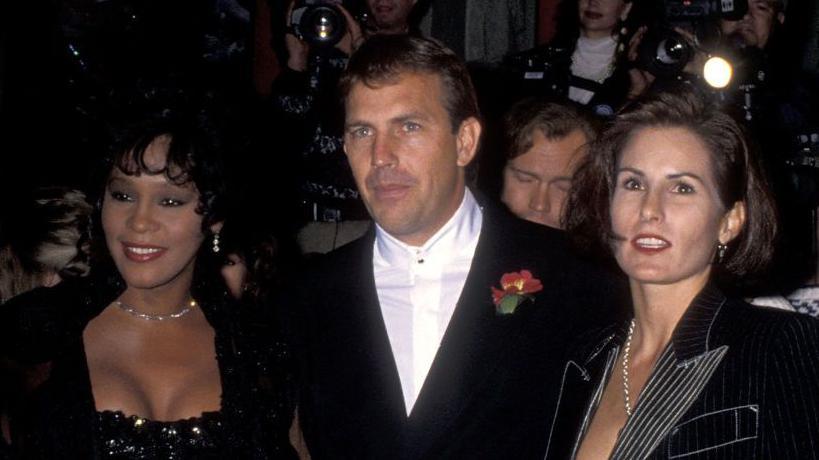 Left to right: Whitney Houston, actor Kevin Costner and his wife Cindy Costner attending The Bodyguard Hollywood premiere in November 1992. They are all dressed in black  formal wear.