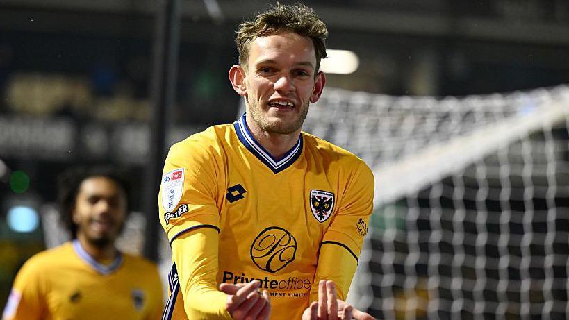 Footballer Callum Maycock playing football for AFC Wimbledon, raising his hands and fingers and smiling to celebrate a goal in a yellow shirt with dark blue trim with a goal and a team-mate blurred behind him