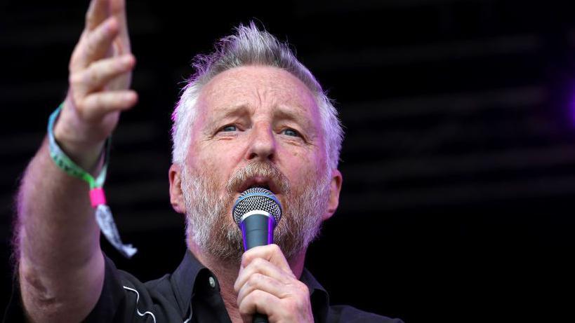 Billy Bragg on stage holding a microphone in one hand and his other hand raised. He has white hair and a brown beard.