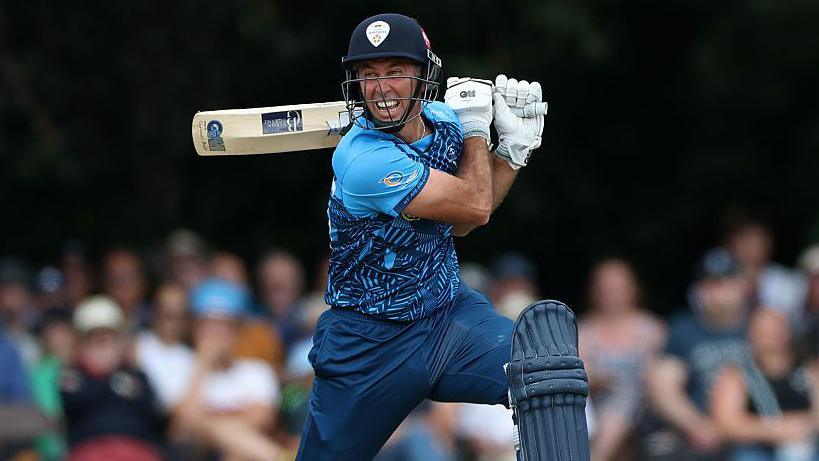 Wayne Madsen in blue Derbyshire Falcons kit holds his bat horizontally behind his head after playing a shot