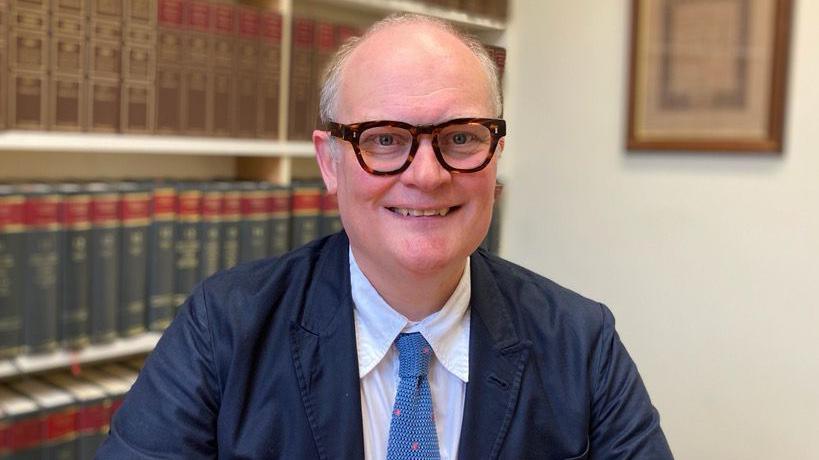 Gary Rycroft wearing thick tortoise shell rimmed glasses and a suit with a baby blue tie in an office with legal books in the background