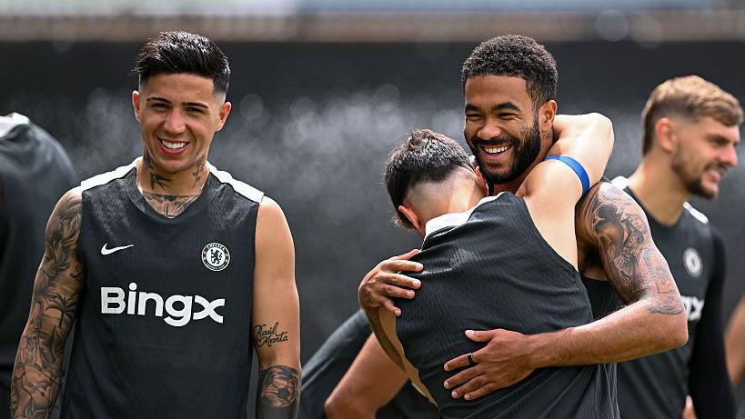 Enzo Fernandez and Reece James wear black Chelsea training vests and grin at the camera as another player with his back to the photographer puts his arms around James' neck