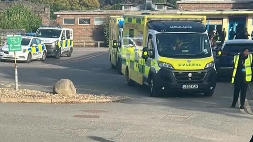 Two ambulances, a police car and police van attend an incident. They are parked in two rows on a concrete car park outside a single-storey building. A woman wearing a high vis vest and blue lanyard stands on the right of the shot, and there are five or six people in the distance, milling around in front of the building.