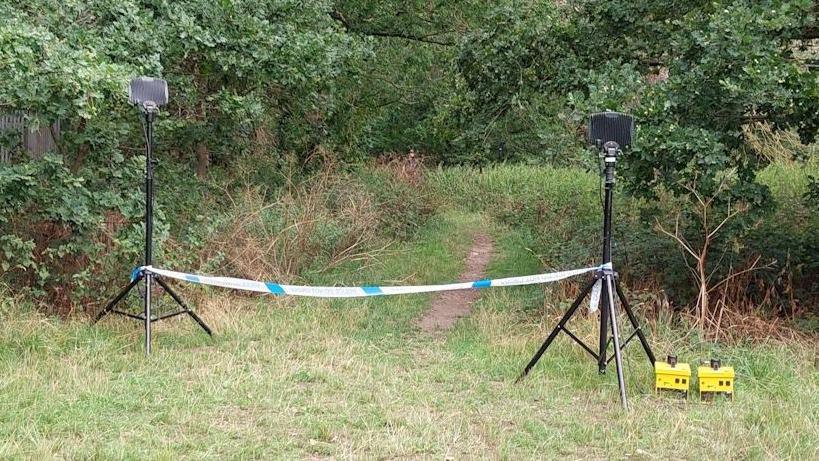 A police cordon in place in a field, with trees beyond it.