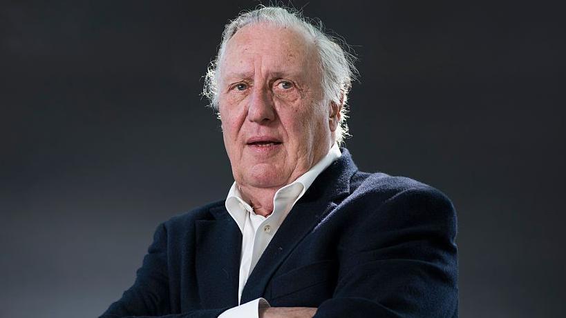 Frederick Forsyth is seen in later life.  He is looking slightly off camera in a head and shoulders portrait. His arms are folded and he is wearing a white shirt and blue jacket.
