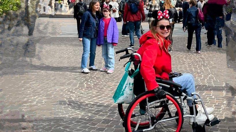 Amy in a red coat and wearing a Minnie Mouse head piece while sat in her wheelchair at Disney Land