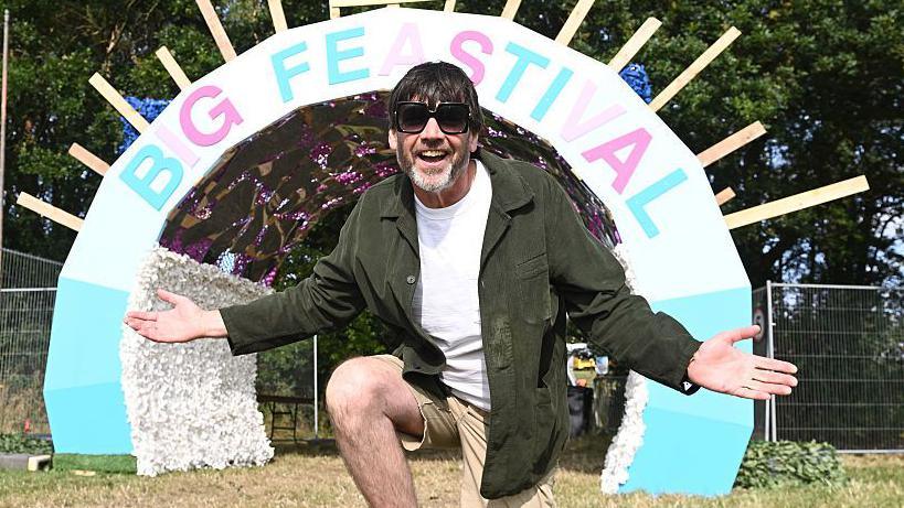 Alex James, wearing sunglasses, a green shirt and khaki shorts, on one knee with arms open, in front of the Big Feastival entrance