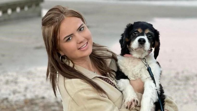 A young woman in a white jacket holding a black and white dog next to her. She is looking into the camera and smiling in front of the sea