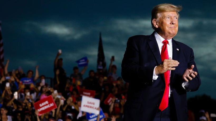 Donald Trump claps as a crowd of supporters cheer behind him.