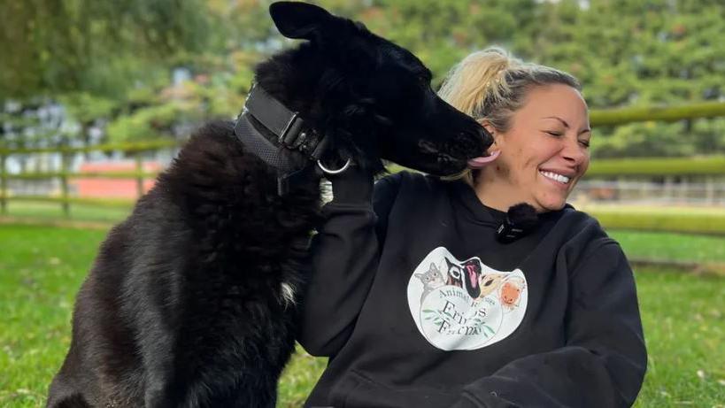 Jodie Marsh wearing a black hoodie and smiling with her eyes closed as a jet black dog licks her right cheek. Jodie has blonde hair.