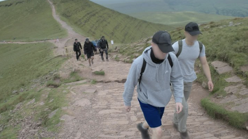 Six Active Club members, wearing sportswear and caps, walk up a stone pavement on the side of a hill, with a green valley stretching out below them