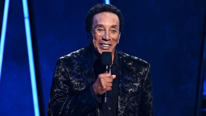 Smokey Robinson at the American Music Awards 50th Anniversary Special at LA Center Studios in Los Angeles, California. He is wearing a black patterned jacket and is holding a microphone in his right hand.