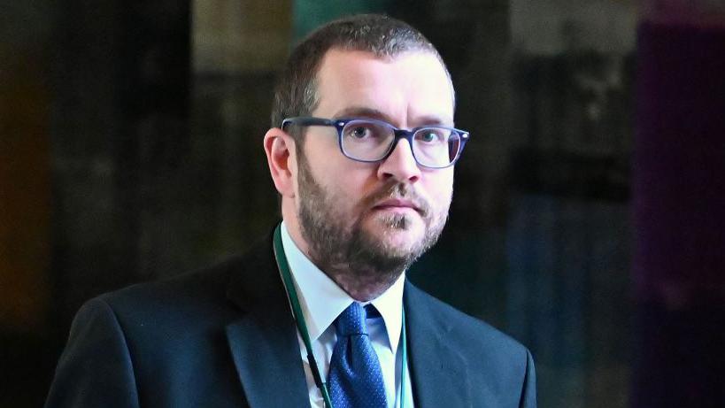 Oliver Mundell with short cut hair and a beard and glasses wears a navy suit with the blue shirt and a blue tie