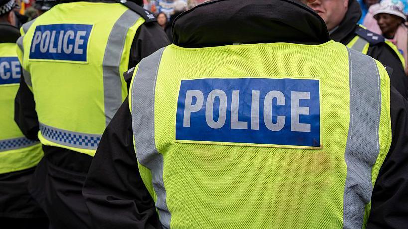 Stock photo of a police man facing away form the camera. He is wearing a black jacket with a yellow high vis jacket that reads POLICE on the back.