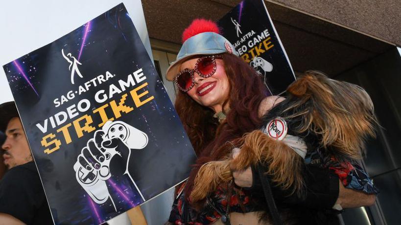 A costumed performer wears a fake fur coat and red-tinted sunglasses as she carries a placard with the words "Sag-Aftra video game strike" and a picture of a hand clutching a gamepad printed on it.