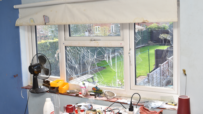 A smashed window inside the house with chemicals on a table. You can see a garden, with green grass, a fan, and plugs. 