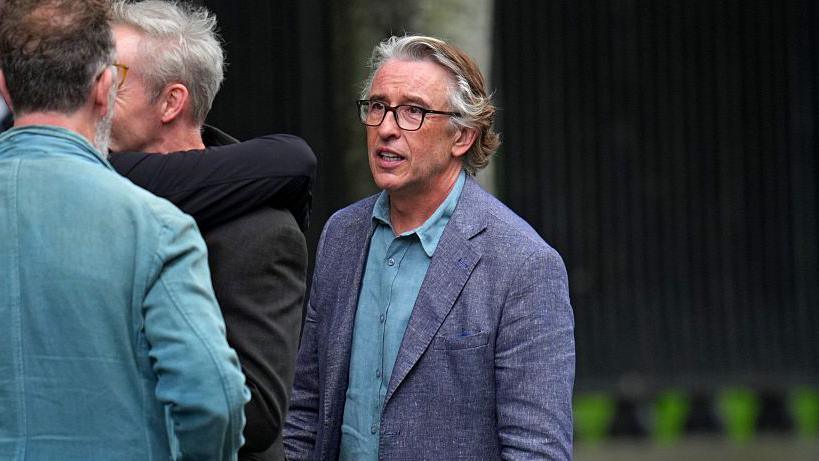 A man with medium-length grey and brown hair stands, looking at another man. He is wearing a pair of glasses, a blue shirt, and a lilac blazer. 