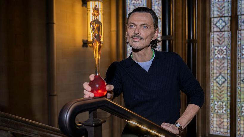 Matthew Williamson, with mid-length dark hair and a goatee beard, wears a dark blue jumper and white T-shirt and holds his Brit Awards design, a yellow-amber statuette of a woman wearing a crested helmet, on the top of a wooden bannister in front of a stone wall and a multi-coloured stained glass window in the University of Manchester