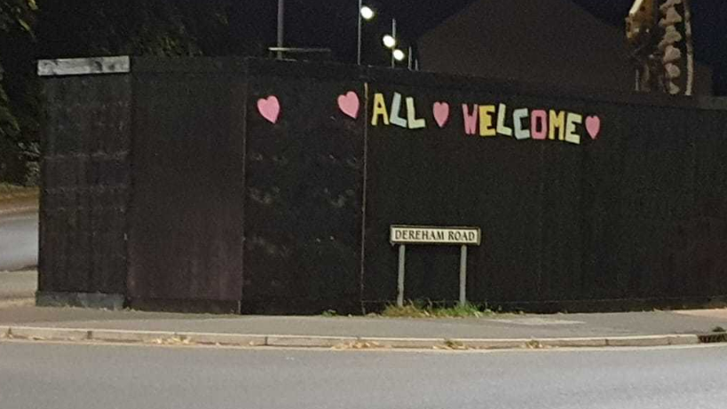 "All welcome" spelled out in what appears to be paper lettering in pastel shades pasted on to black hoarding. It is accompanied by four pink paper hearts. A "Dereham Road" sign is in front of it.