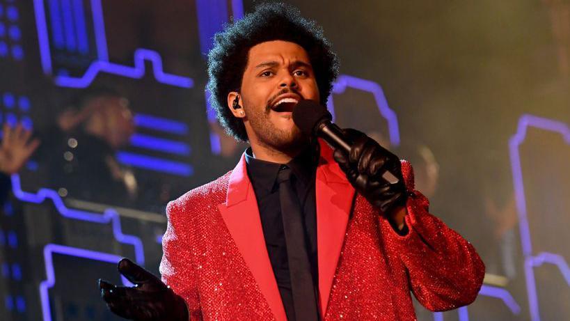 Singer The Weeknd holding a microphone and wearing a bright red jacket