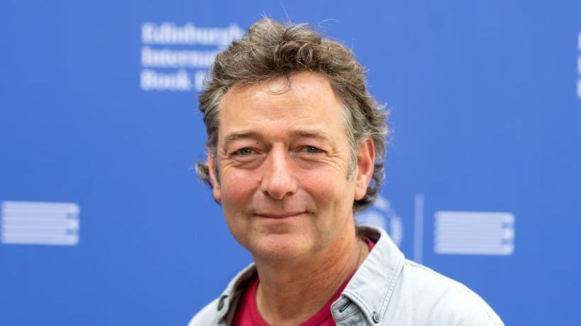 Ned Boulting smiles at the camera during a book festival. He has short brown and grey hair. He is wearing a blue shirt with a red t-shirt underneath. He is standing in front of a blue background.