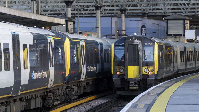 A file image showing two trains side by side as they are pulled into the platform at Waterloo station, both with a white and blue South Western Railway logo on the side of the carriages and yellow, light and dark blue livery