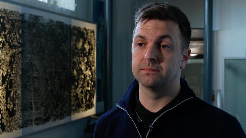 Conchúr Ó Muadaigh - a man with short brown hair, wearing a navy jumper, stood beside a large framed print