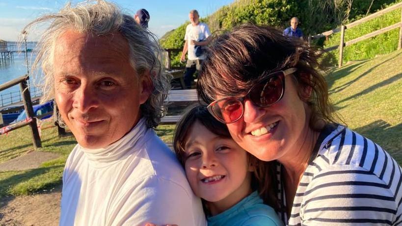 A man, boy and woman smile in the sunshine, sat close together in a row, facing the camera. The man has grey hair to his shoulders and is wearing a white polo neck, behind him the boy is grinning with missing teeth and wearing a pale blue T-shirt. Behind the boy the woman has brown hair swept back from her face and sunglasses. She is wearing a black and white striped top.