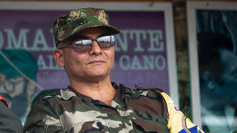 The man known as alias Iván Mordisco speaks during the announcement by the Central General Staff (EMC) armed group to open peace talks with the Colombian government on 16 April, 2023. He wearing a camouflage shirt and an armband in the colours of the Colombian flag. He is also wearing a camouflage cap and dark glasses. He is holding some sheets of paper in his hands.