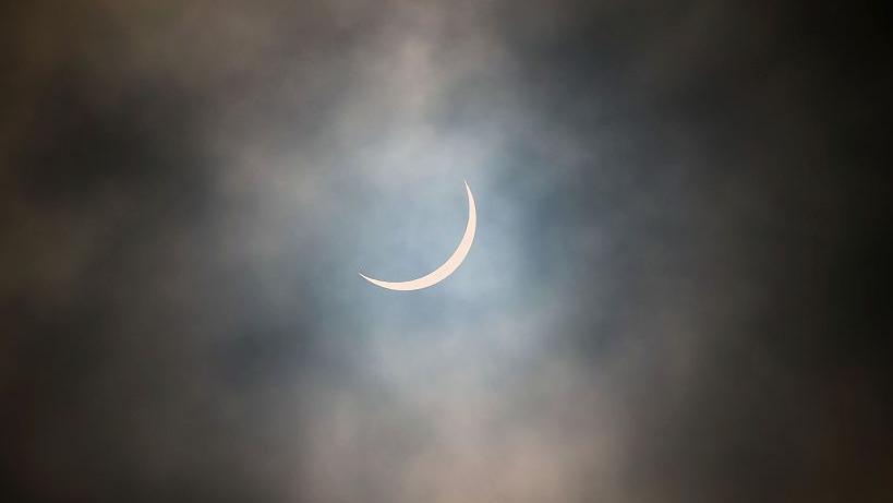 The sun is pictured during a partial solar eclipse. The sky is dark and cloudy. The sun is a thin arc in the sky.