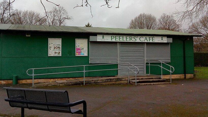 A green cafe in a Bradford park, with "Peelers' Cafe" written in green letters on the sign. Grey shutters are down over the door and windows. A park bench is in the foreground. 