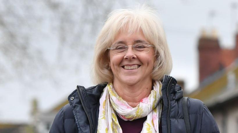 A woman with bobbed light blonde hair and glasses, wearing a navy puffer jacket and white and pink floral scarf and smiling at the camera.