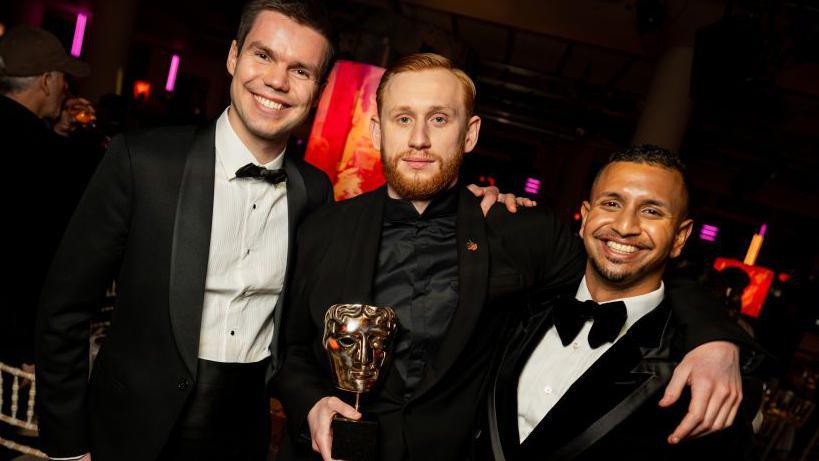 Franz Böhm, Oleksandr Rudynskyy and Hayder Rothschild Hoozeer are dressed in suits and smiling while holding a golden bafta trophy