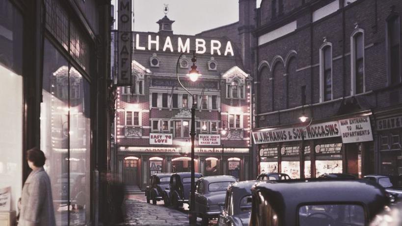 An old photograph of the street with the Alhambra theatre at the end, old fashioned cars line the street