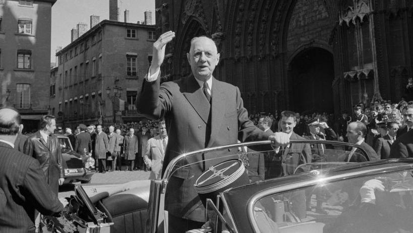 General de Gaulle is seen in a black and white photo in civilian dress. He is standing in the back of an open top car waving at the crowd.