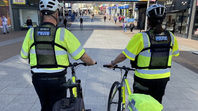 The back of two police officers, who are both wearing high-visibility neon jackets, walking down a high street with their bikes. There are a number of people outside of shops in the high street. Both officers are wearing helmets.