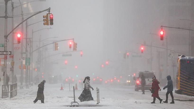 People walking across Sixth Avenue as snow falls. There's snow on the ground and lots of traffic lights on red. Cars are in the distance with their brake lights on.