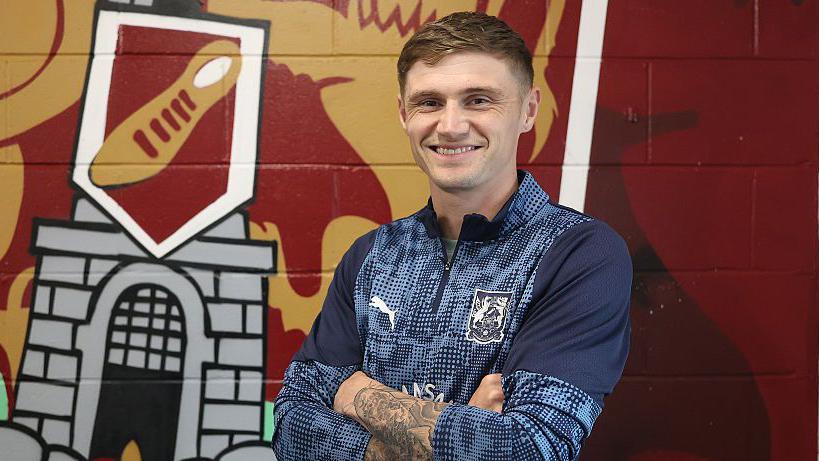 Northampton Saints striker Harvey Saunders folding his arms in front of the club crest on the wall after signing.