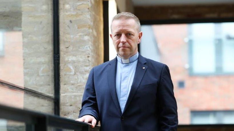 Andrew is wearing a minister's collar and a blue shirt, navy suit and he is standing by a window looking at the camera. He has blond short hair and blue eyes. 