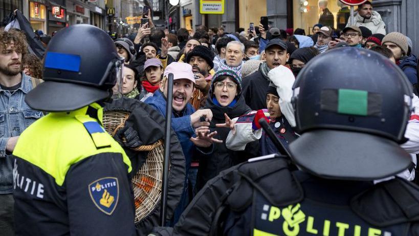 Police officers in riot gear face off with protesters who seem to be yelling or chanting.