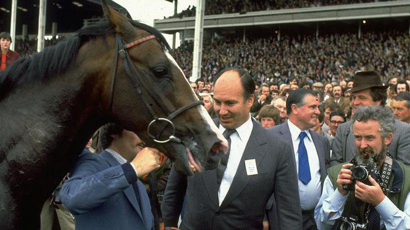 A brown horse wearing a bridle stands to the left of the image and a balding man wearing a grey suit is looking at the horse and smiling. Standing beside him is a small man with grey curly hair holding a camera. There is a crowd of spectators in the background.