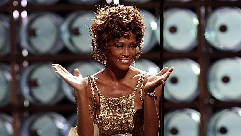 Whitney Houston performing on stage during the 2004 World Music Awards at the Thomas and Mack Center, Las Vegas in 2004. She is wearing a gold strappy dress.