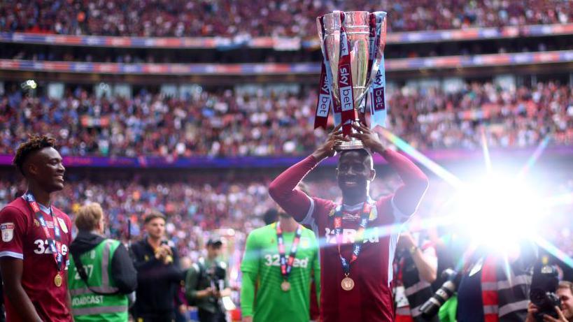 Adomah stands on Wembley pitch holding the Skybet Championship play-off final trophy aloft with a camera flash shining over his left shoulder