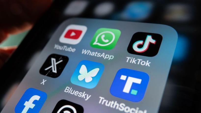 An iPhone screen is lit up and shows an open folder with various social media logo apps including YouTube, WhatsApp, TikTok, X, Bluesky, TruthSocial and Facebook