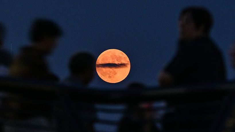 People are seen as shadows. In the background a red hued 'Blood Moon' in the sky. 