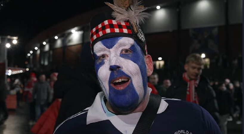 A man wears a Scotland top and checked headband with a blue and white saltire painted on his face. He is outside Hampden Park and it is dark