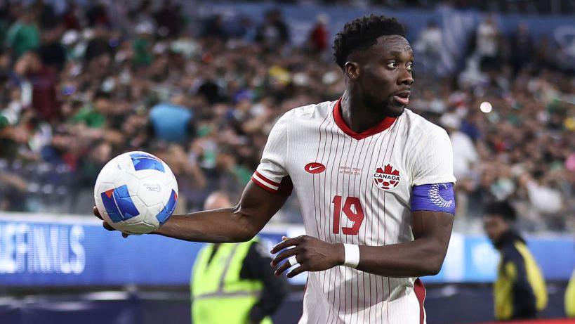 Alphonso Davies holding a ball while in action for Canada, wearing the captain's armband