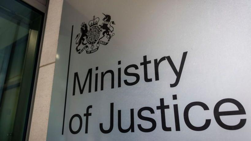 'Ministry of Justice' entrance sign with emblem, a lion and a seahorse with crowns and horns and Latin that is too small to read. 

The writing is black and the background is white. 